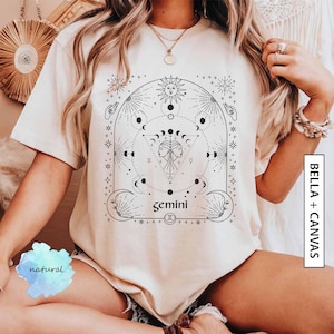 May include: A white t-shirt with a black graphic design featuring a celestial illustration with the zodiac sign Gemini. The design includes a sun, moon, stars, and a triangle with two faces inside. The text "gemini" is written below the triangle.