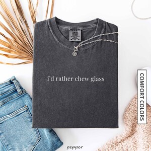 May include: A dark gray t-shirt with the text "i'd rather chew glass" in white. A silver necklace with a pendant is draped across the shirt. The shirt is folded and placed on a white surface next to a pair of blue jeans.