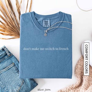 a blue t-shirt with the text "don't make me switch to french" printed on it, along with a denim jacket, jeans, and a beige sweater.