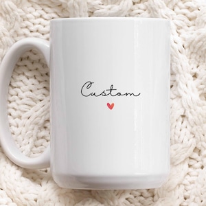 Può includere: Tazza in ceramica bianca con un manico grande. La parola "Custom" è scritta in corsivo nero, con un piccolo cuore rosso sotto. La tazza è posta su uno sfondo bianco strutturato.
