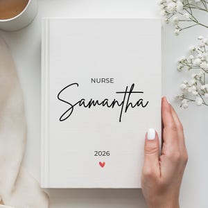 Könnte beinhalten: Ein weißes Notizbuch mit dem Namen "Samantha" in eleganter Schrift, dem Wort "NURSE" darüber und dem Jahr "2026" darunter, mit einem kleinen roten Herz. Das Notizbuch wird von einer Person gehalten.