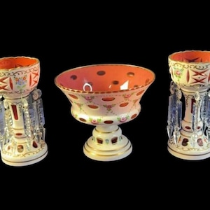 Może przedstawiać: Trzy antyczne szklane misy i cokoły. Misy mają czerwone wnętrze i są zdobione wzorami kwiatowymi i geometrycznymi. Cokoły posiadają kryształowe akcenty oraz biało-złotą podstawę. Tło jest czarne.