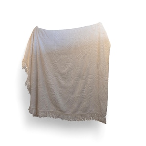 May include: Cream-coloured, textured throw blanket with a detailed pattern and fringe trim. The blanket is draped, showcasing the intricate design and soft texture. The fringe adds a decorative element to the bottom edge.