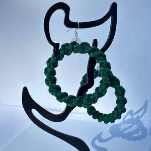 May include: A pair of dark green macrame earrings displayed on a black metal earring stand. The earrings are circular, with a woven texture. The stand has a curved design, and the background is a light blue.
