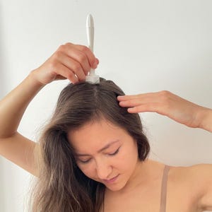 Puede incluir: Una persona utiliza un aplicador blanco para el cuidado del cabello en su cabello. El aplicador tiene un mango largo y una punta redondeada. La persona sostiene el aplicador con una mano y usa la otra para guiarlo.