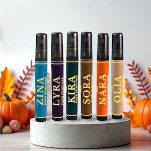 Puede incluir: Seis frascos de perfume para el cabello con tapas negras y etiquetas coloridas. Las etiquetas muestran nombres como ZINA, LYRA y NARA, con el texto "HAIR PERFUME". Los frascos están dispuestos sobre una plataforma gris y circular, con calabazas y hojas de otoño en el fondo.