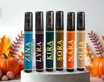 May include: Six hair perfume bottles with black caps and colorful labels. The labels display names like ZINA, LYRA, and NARA, with "HAIR PERFUME" text. The bottles are arranged on a gray, circular platform, with pumpkins and fall foliage in the background.
