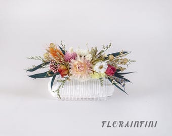 Puede incluir: Un peine para el cabello adornado con flores secas en tonos rosa, naranja, blanco y rojo. El arreglo floral se encuentra sobre un peine blanco, complementado con hojas verdes oscuras y hierba seca. El texto "FLORAINFINI" es visible en la parte inferior.
