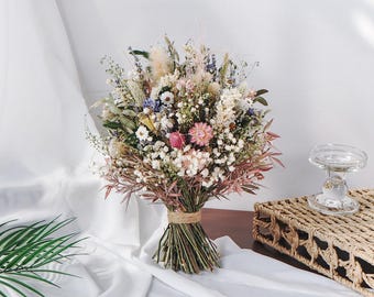 May include: A vibrant bouquet of dried flowers, featuring a mix of pink, white, and purple blooms, tied with twine. The arrangement includes various textures and shapes, creating a natural and rustic aesthetic. The bouquet is set against a white backdrop.
