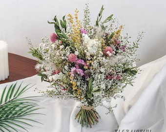 May include: A vibrant bouquet of dried flowers, featuring a mix of white, yellow, pink, and green hues. The arrangement is tied with twine and rests on a white cloth. The bouquet includes various textures and shapes, creating a natural and rustic aesthetic.
