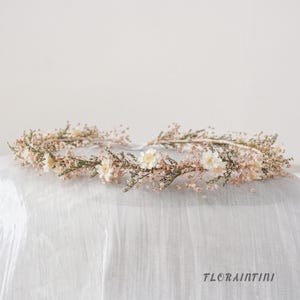 Puede incluir: Una corona de flores con pequeñas flores blancas similares a las margaritas, intercaladas con delicado follaje seco en tonos rosa claro y verde. El tocado circular está construido con tallos finos y flexibles e incluye el texto FLORAINFINI.