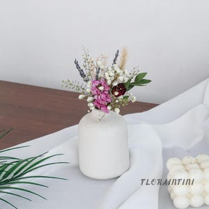 May include: A small, white ceramic vase filled with a colorful arrangement of dried flowers. The bouquet includes pink, white, and purple blooms, with green foliage. The vase sits on a white cloth, with the text "FLORAINTINI" visible.