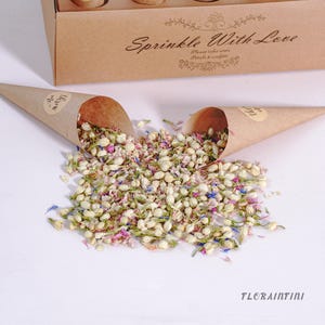 Op de afbeelding: Een close-up van gedroogde bloemblaadjes en confetti die uit twee bruine papieren kegels vallen. De kegels staan voor een bruine doos met de tekst "Sprinkle With Love". De bloemblaadjes zijn wit, roze en blauw.