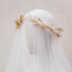 May include: A bridal headpiece with a crown of dried flowers and wheat, and small white beads. A sheer white veil drapes down, creating a soft, ethereal look. The headpiece is set against a neutral background.