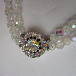 May include: A double strand necklace made of clear iridescent glass beads with a silver clasp featuring a large iridescent rhinestone in the centre.