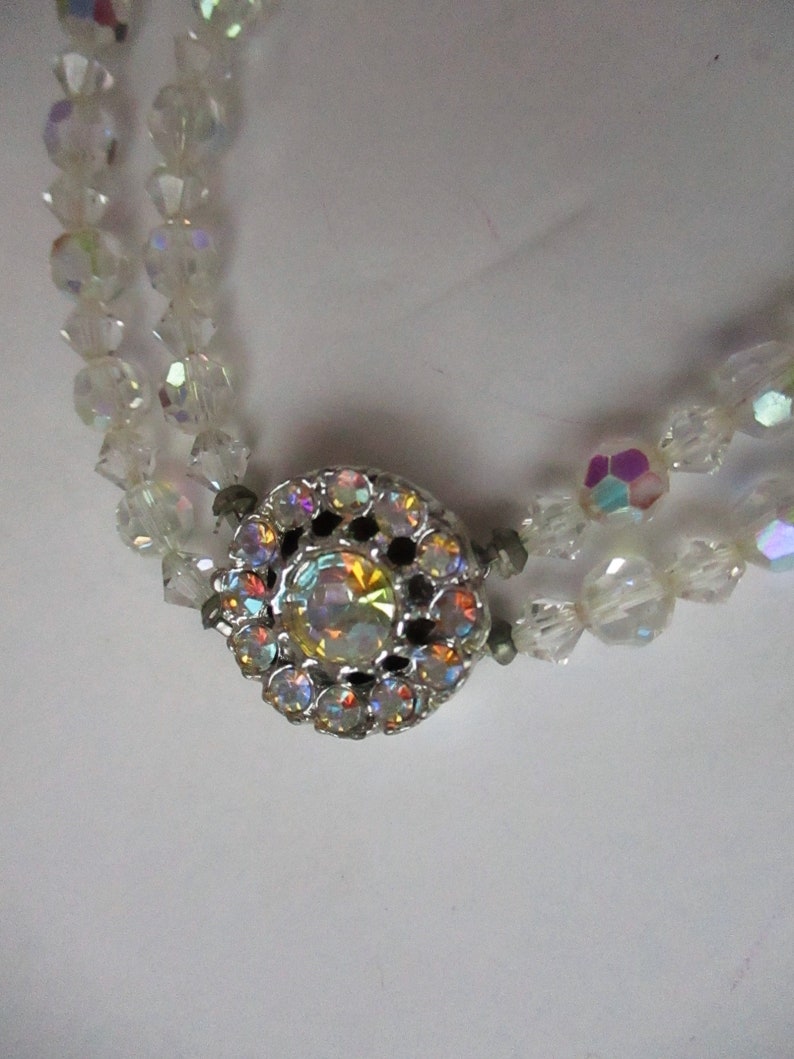 May include: A double strand necklace made of clear iridescent glass beads with a silver clasp featuring a large iridescent rhinestone in the centre.