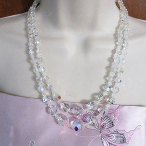 May include: A double strand iridescent crystal necklace with clear faceted beads. The necklace is on a white mannequin bust and a pink fabric background with a butterfly design.