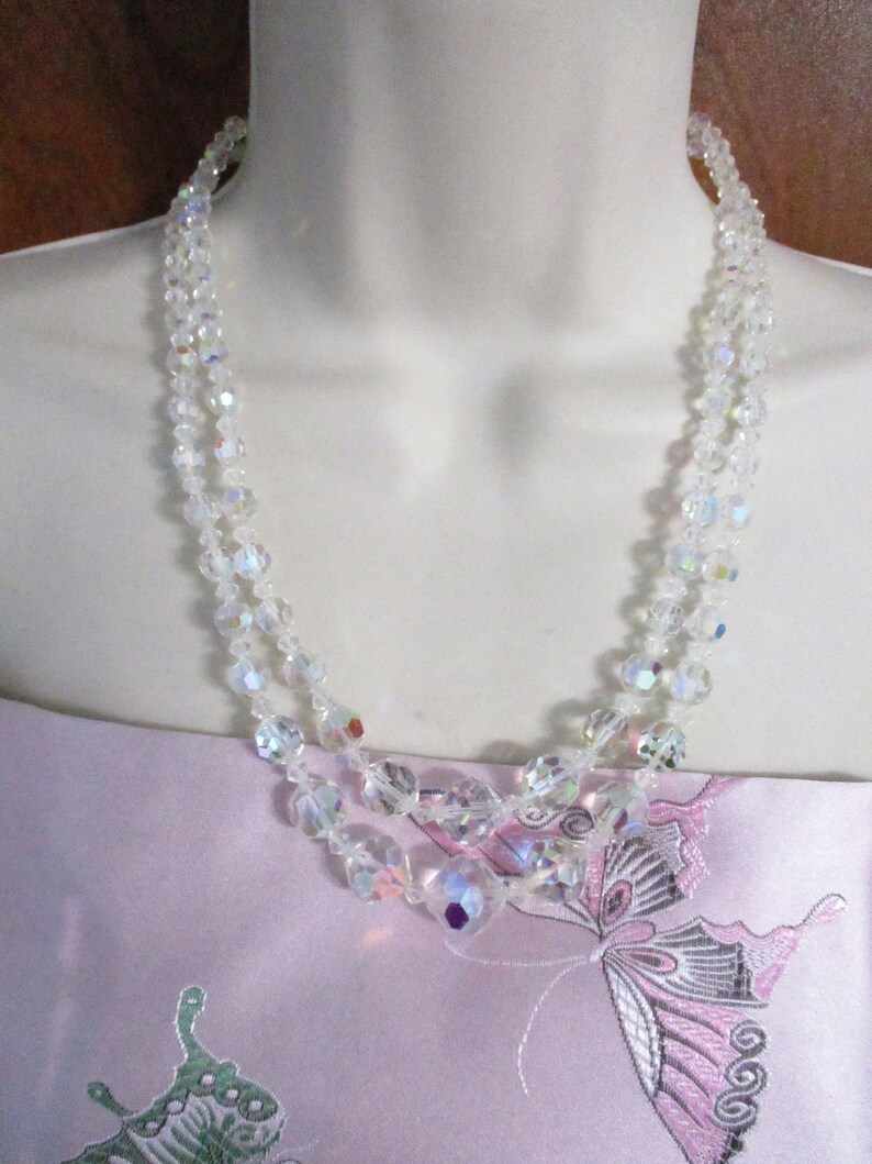 May include: A double strand iridescent crystal necklace with clear faceted beads. The necklace is on a white mannequin bust and a pink fabric background with a butterfly design.