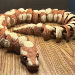 May include: A segmented toy snake in shades of brown and cream, with a textured, scaly appearance. The snake's head is detailed with eyes and a snout. The toy is posed on a wooden surface.