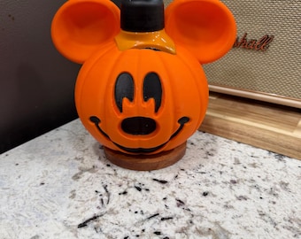 May include: An orange soap dispenser shaped like a pumpkin with round ears and a black pump mechanism. The pumpkin features a carved-style face with black eyes, a nose, and a wide smile. The dispenser sits on a small, circular wooden base.