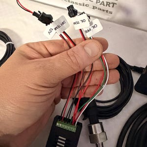 May include: A black electronic component with various coloured wires and connectors. The wires are red, black, white, and green. Labels read "DIFF TEMI 12V", "OIL TEM" and "OIL PRE". The image also shows a pressure sensor and coiled black cables.
