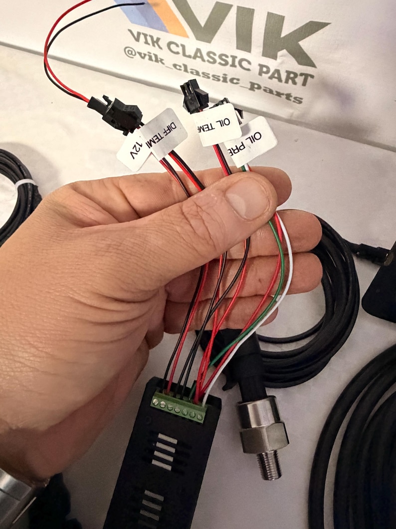 May include: A black electronic component with various coloured wires and connectors. The wires are red, black, white, and green. Labels read "DIFF TEMI 12V", "OIL TEM" and "OIL PRE". The image also shows a pressure sensor and coiled black cables.