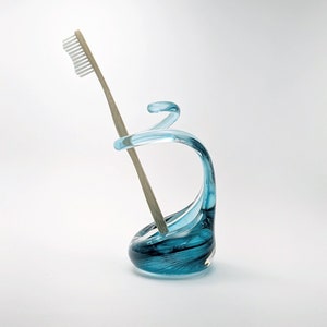 May include: A teal blue glass toothbrush holder with a swirling design, holding a bamboo toothbrush with white bristles. The holder has a unique, artistic shape and sits on a white surface.