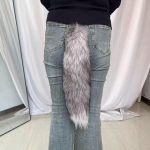 Puede incluir: Una cola de piel sintética plateada y blanca sujeta a la parte trasera de unos vaqueros azules. La cola es larga y esponjosa, con un degradado de colores de plata a blanco. Los vaqueros son de mezclilla de lavado claro.