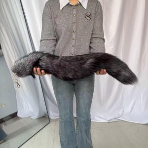 May include: A long, faux fur tail in shades of black and silver, held horizontally. The tail has a fluffy texture and a keychain attachment. The item is designed to be worn as an accessory.