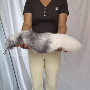 May include: A long, fluffy faux fur tail accessory featuring a gradient of colors transitioning from dark gray and black at the base to a lighter gray and white toward the tip. The tail includes a small gold-toned metal lobster clasp attachment.