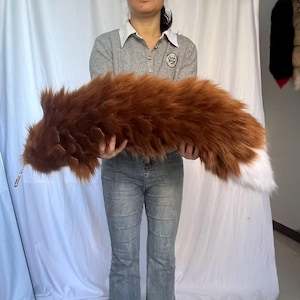May include: A long, plush, faux fur tail accessory in a rich reddish-brown color with a distinct white tip. The tail features a soft, shaggy texture and includes a small metal clasp attached to one end for secure fastening or display.