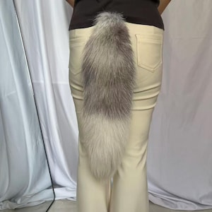 May include: A long, fluffy, faux fur animal tail in shades of grey, white, and brown, attached to the back of a pair of cream-colored trousers. The tail features a gradient of dark grey at the top transitioning to a lighter, creamy white at the tip.