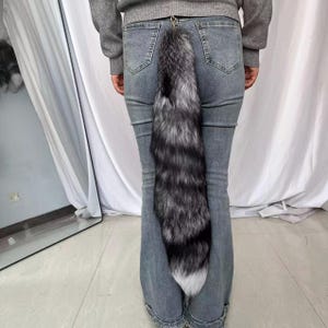 Może przedstawiać: Srebrno-czarny ogon z imitacji futra z białą końcówką, przymocowany do srebrnego metalowego zapięcia. Ogon zwisa z tyłu osoby noszącej szare dżinsy.