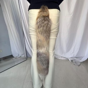 May include: A long, fluffy tail accessory with a mix of brown, gray, and cream-colored fur. The tail is attached to the back of a pair of cream-colored pants. The fur appears soft and natural, with varying shades creating a textured look.