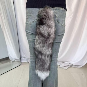 Puede incluir: Una cola de piel sintética, principalmente plateada y negra, sujeta a la parte trasera de unos vaqueros grises. La cola tiene una textura esponjosa y una punta blanca. La cola está sujeta con un cierre dorado.