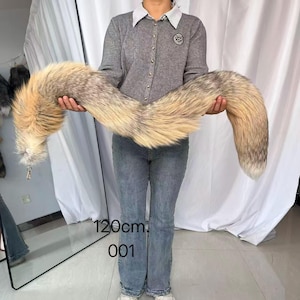 Peut inclure: Une longue et duveteuse queue de renard aux tons beige, crème et marron est présentée. La queue mesure environ 120 cm de long, avec un porte-clés attaché à la base. Le numéro "001" est imprimé sur l'image.