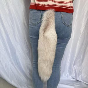 Könnte beinhalten: Ein langer, flauschiger, cremefarbener Kunstfellschwanz, der an der Rückseite einer hellblauen Jeans befestigt ist. Der Schwanz hat eine konische Form und ein weiches, strukturiertes Aussehen. Die Person trägt ein rot-weiß gestreiftes Oberteil.