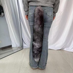 Könnte beinhalten: Ein langer Kunstfellschwanz in Grau- und Silbertönen, der an der Rückseite einer Jeans befestigt ist. Der Schwanz ist dick und flauschig, mit einer leichten Krümmung. Die Person trägt einen grauen Pullover.