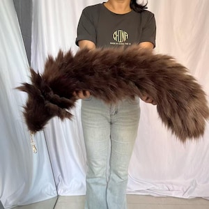 May include: A long, dark brown faux fur tail accessory featuring a gold-toned metal lobster clasp at one end. The plush, textured material is thick and voluminous, extending horizontally across the frame with a tapered, pointed tip.