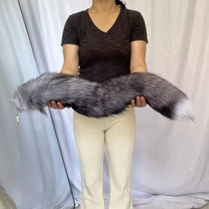 Può includere: Una lunga coda in pelliccia sintetica morbida nei toni dell'argento, del grigio e del nero. La coda presenta un aspetto spesso e strutturato con una sfumatura di pelo grigio scuro e nero che sfuma verso una punta bianca. Un clip metallico è fissato.