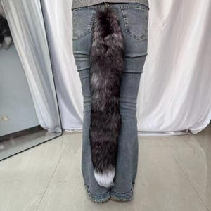 Może przedstawiać: Długi, sztuczny ogon z futra w odcieniach szarości i czerni, z białą końcówką, przymocowany do pary szarych dżinsów. Ogon jest puszysty i wydaje się być wykonany z miękkiego materiału. Ogon jest przymocowany do dżinsów metalową klamrą.