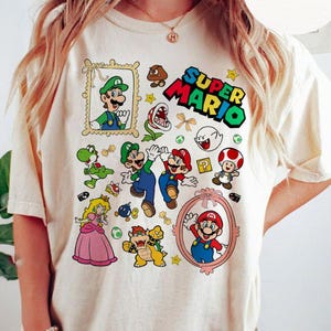 May include: A cream colored t-shirt with a graphic print of various Super Mario characters, including Mario, Luigi, Yoshi, and Toad. The text "SUPER MARIO" is printed in red and black letters.