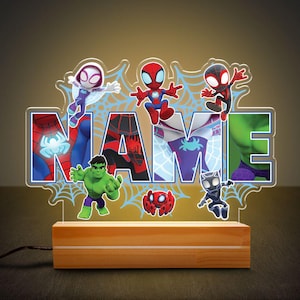 May include: A light-up name sign with a Spiderman theme. The sign is made of clear acrylic and features various Spiderman characters, including Spiderman, Green Goblin, and Spider-Gwen. The sign is illuminated by LED lights and has a wooden base.