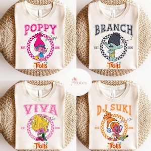 May include: Four cream-colored t-shirts with different Trolls character designs. Each shirt features a character name in a unique font, a cartoon illustration, and the word "Trolls". The shirts are on a woven surface.