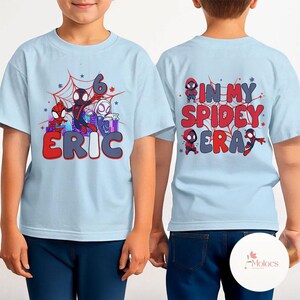 May include: Two light blue t-shirts with a Spiderman theme. The front shirt has a graphic of Spiderman characters and the text "6 ERIC". The back shirt has a graphic of Spiderman characters and the text "IN MY SPIDEY ERA".
