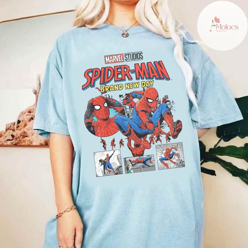 Op de afbeelding: Lichtblauw T-shirt met een Spider-Man-afbeelding. Het ontwerp bevat de tekst "MARVEL STUDIOS SPIDER-MAN BRAND NEW DAY" in rood en geel. De afbeelding toont Spider-Man in verschillende actieposes, met stripboekstijl panelen.