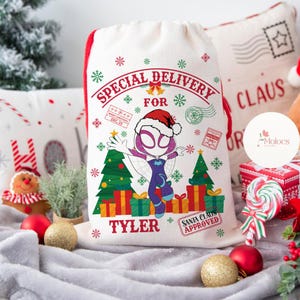 May include: A white Christmas gift bag with a red drawstring, featuring a graphic of a superhero in a Santa hat, Christmas trees, and wrapped gifts. Text includes "SPECIAL DELIVERY FOR TYLER" and "SANTA CLAUS APPROVED."