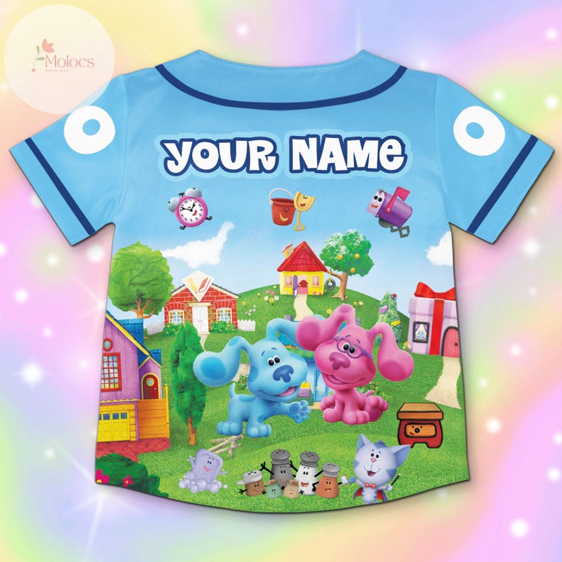 May include: Light blue baseball jersey featuring a colorful cartoon scene with the text "YOUR NAME". The design includes cartoon dogs, houses, and other animated characters. The jersey has blue trim and white accents.