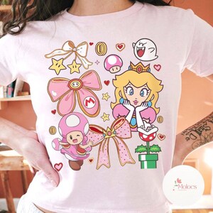 Puede incluir: Camiseta rosa claro con un gr&aacute;fico colorido de la Princesa Peach, Toad y otros personajes de la serie de videojuegos Super Mario. El dise&ntilde;o incluye lazos, estrellas, monedas y corazones. La camiseta es de cuello redondo.