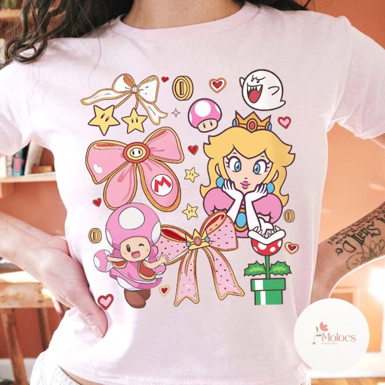 Puede incluir: Camiseta rosa claro con un gr&aacute;fico colorido de la Princesa Peach, Toad y otros personajes de la serie de videojuegos Super Mario. El dise&ntilde;o incluye lazos, estrellas, monedas y corazones. La camiseta es de cuello redondo.